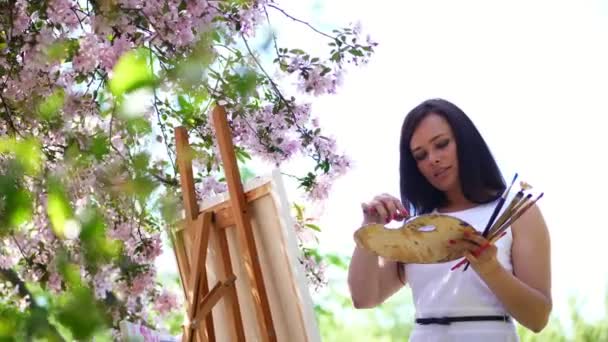 Een mooie vrouw schilder in witte jurk, tijdens de bloei voorjaar appelboomgaard, ze verf mengt met een penseel op de pallet. de wind zwieren bloeiende takken over ezel met doek, — Stockvideo