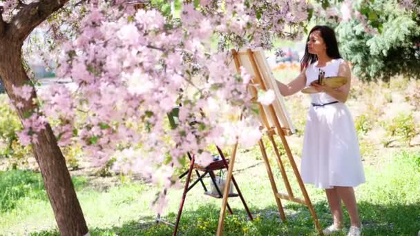 En vacker kvinna målare i vit klänning, konstnär målar en bild av blommor i blommande våren äppelträdgård, håller hon en palett med färger och en pensel i händerna — Stockvideo