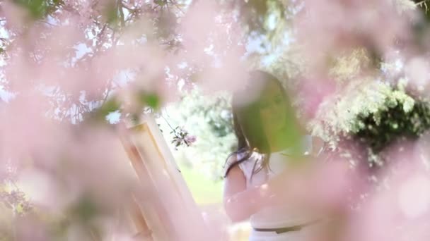 Una hermosa pintora en vestido blanco, artista pinta un cuadro de flores en floreciente huerto de manzanas de primavera, ella sostiene una paleta con pinturas y un pincel en sus manos — Vídeos de Stock