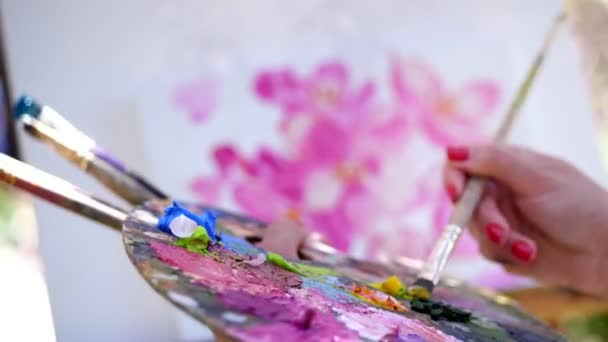 Gros plan, mains féminines, peintre, artiste peint un tableau de fleurs, elle tient la palette avec des peintures et des pinceaux, mélange des peintures avec un pinceau sur la palette — Video