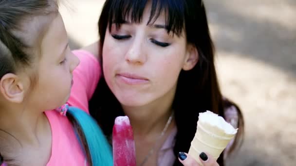 Bella ragazza di otto anni, bionda, con lentiggini, e sua madre, una donna bruna, mangiare gelato bianco in tazza di waffle, gelato rosa su bastone, trattare, nutrirsi a vicenda. ridere, divertirsi . — Video Stock