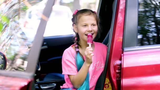 Retrato, chica bonita de ocho años, rubia, con pecas, y coletas multicolores, comiendo helado rosa en un palo, sonriendo. se sienta en el coche, cerca de la puerta abierta — Vídeo de stock