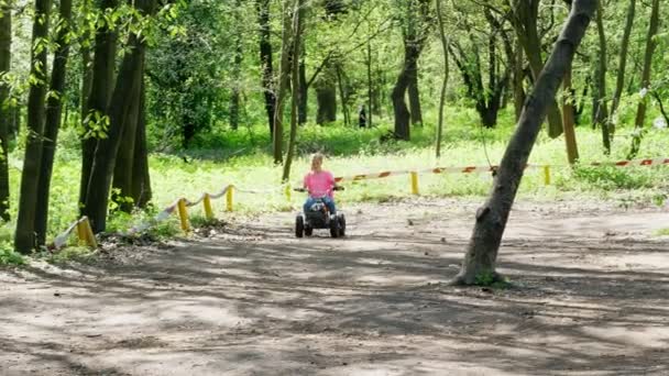 Zomerdag in openbare pine stadspark, gekleurde klein meisje in een helder roze T-shirt met pigtails rijden, rijden op een childs vier wiel motorfiets, quad mini moto, vierwieler. Kinderen zomervakantie — Stockvideo