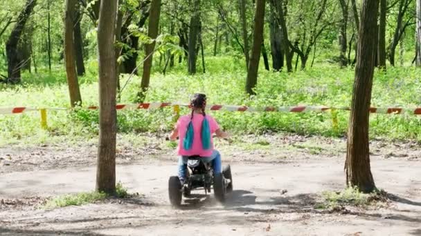 Sommardag i offentliga pine city park, färgade liten flicka i en ljust rosa T-shirt med flätor rider, kör på en childs fyra hjul motorcykel, mini moto quad, fyrhjuling. Barnen sommarlov — Stockvideo