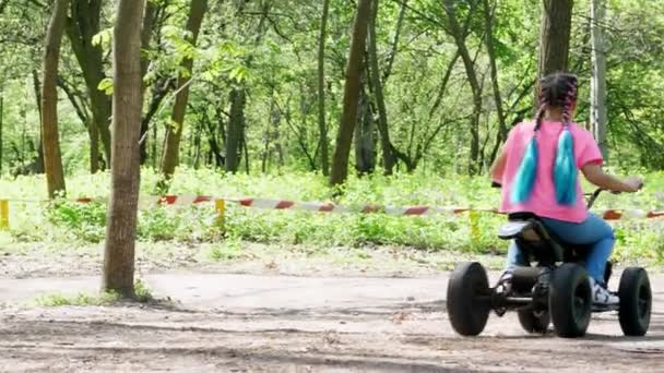 Літній день у громадському сосновому міському парку, маленька дівчинка в яскраво-рожевій футболці з кольоровими косичками, їзда на дитячому чотириколісному мотоциклі, міні мото квадроциклі. Дитячі літні канікули — стокове відео