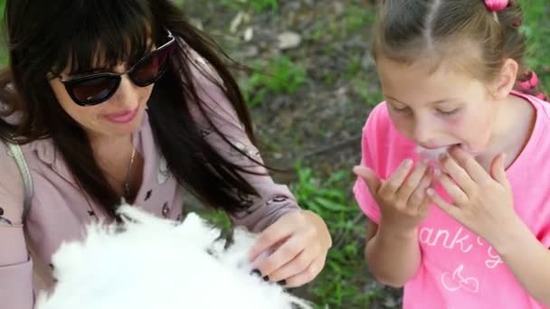 Donna bruna, indossa gli occhiali da sole, con sua figlia, ragazza di 8 anni, con le trecce blu, mangiare zucchero filato dolce, filo interdentale, ridere, divertirsi, nel parco della città, in estate — Video Stock