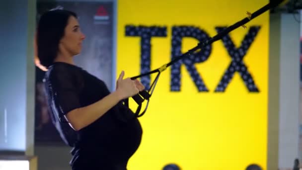 Cherkasy, Oekraïne, 20 november 2019: Trx-training. jonge zwangere atletische vrouw, met een grote buik, in een zwart nauwsluitende trainingspak, doet oefeningen met trx fitness riemen in de sportschool — Stockvideo