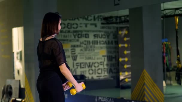 Jonge zwangere atletische vrouw, met een grote buik, in een zwart trainingspak, doet oefeningen met halters voor armspieren in de sportschool — Stockvideo