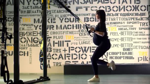 CHERKASY, UCRANIA, 20 de noviembre de 2019: Entrenamiento TRX. mujer atlética embarazada joven, con un vientre grande, en un chándal ajustado negro, está haciendo ejercicios con correas de fitness trx en el gimnasio — Vídeos de Stock