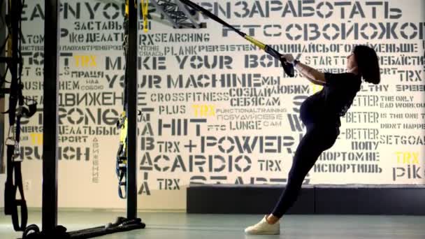 ЧЕРКАСИ (УКРАИНА), 20 ноября. молодая беременная спортсменка, с большим животом, в черном обтягивающем спортивном костюме, делает упражнения с трх фитнес-ремнями в тренажерном зале — стоковое видео