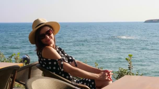 Chica morena en un sombrero y gafas de sol, se sienta en una mesa en un café, en la orilla del mar, admira el paisaje marino. caliente día de verano . — Vídeos de Stock