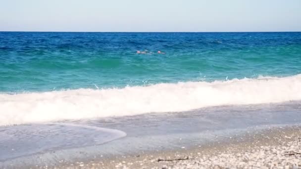 In het turquoise, blauwe water van de zee zwemmen twee mannen. Zomer hete zonnige dag. zachte golven op zee, op het strand. golven met wit schuim worden aangespoeld. — Stockvideo