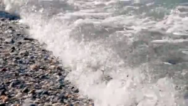 Yakın plan. Yumuşak dalgalar, beyaz köpükle kıyıya vurur, çakıl taşı sahilinde. Akdeniz kıyılarında, sahilde çok renkli yuvarlak deniz çakıl taşları. Sıcak yaz günü. Yunanistan — Stok video