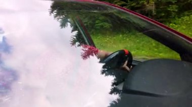 Araba yolcularının ön camından dışarıyı görebiliyoruz. Adam sürüyor. Ağaçların tepeleri ön cama yansıyor. Araba hareket halinde..
