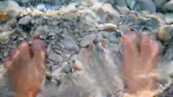 Close-up. menselijke benen in het water. onder het heldere water van de Egeïsche Zee zijn kleine veelkleurige kiezelsteentjes zichtbaar. zonlicht, stralen worden gereflecteerd in water. — Stockvideo