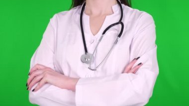 yakın plan, kadın doktor, cerrah, beyaz tıbbi üniforma giymiş ve steteskoplu, yeşil arka planda çapraz ellerle duran.