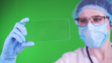 green background. close-up, doctor dressed in medical cap, mask, blue medical gloves, holds a glass card on which it is possible to place an advertisement, text or video.