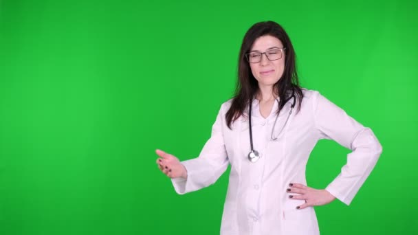 Porträt einer glücklichen jungen Ärztin mit Brille, in weißer Arztuniform und mit Stethoskop, präsentiert, zeigt Kopierraum für Produkt, Text auf grünem Hintergrund. — Stockvideo