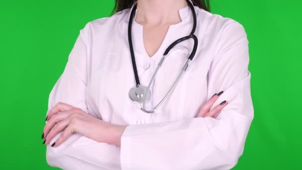 Close-up, médica, cirurgiã, vestida com um uniforme médico branco e com um estetoscópio, de pé com as mãos cruzadas sobre um fundo verde . — Vídeo de Stock