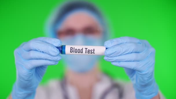 Zelené pozadí. zblízka, lékaři ruce v modrých lékařských rukavicích drží zkumavku s nápisem krevní test. doktor v lékařské čepici, maska. koncepce laboratorního výzkumu. — Stock video