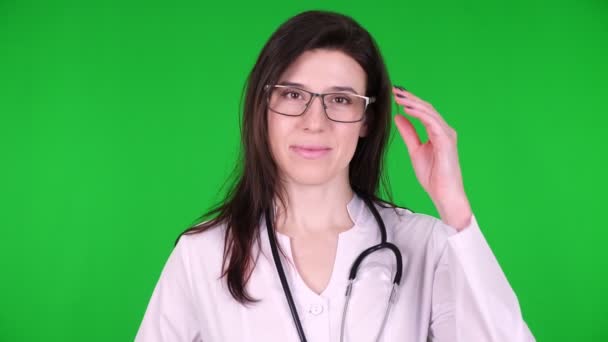 Retrato de jovem médica feliz, cirurgiã de óculos, vestida com um uniforme médico branco e com um estetoscópio. sobre um fundo verde . — Vídeo de Stock