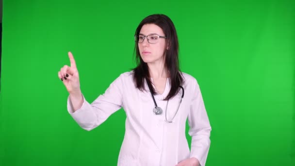 Portret van jonge vrouwelijke arts in bril, gekleed in wit medisch uniform en met stethoscoop, wijzend met de vinger in de ruimte voor product of tekst op groene achtergrond. — Stockvideo