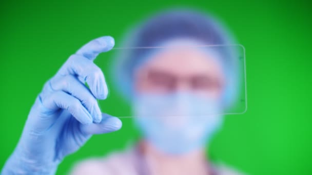 Green background. close-up, doctor dressed in medical cap, mask, blue medical gloves, holds a glass card on which it is possible to place an advertisement, text or video. — Stock video