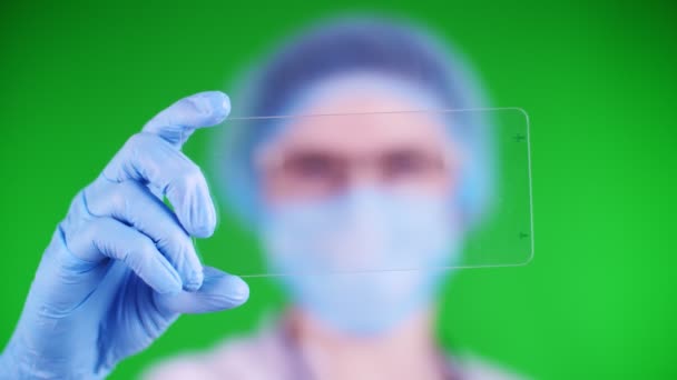 Green background. close-up, doctor dressed in medical cap, mask, blue medical gloves, holds a glass card on which it is possible to place an advertisement, text or video. — Wideo stockowe