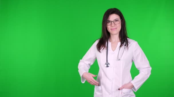 Retrato de la joven médica feliz en gafas, vestida con uniforme médico blanco y con estetoscopio, presentando, mostrando espacio de copia para el producto, texto sobre fondo verde . — Vídeos de Stock