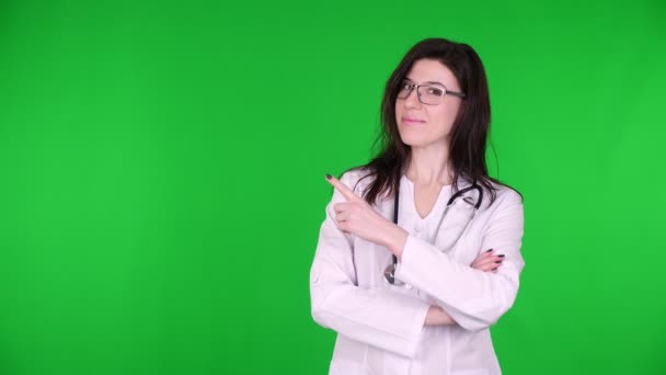 Portret van een vrolijke jonge vrouwelijke arts in bril, gekleed in wit medisch uniform en met stethoscoop, presentatie, met kopieerruimte voor het product, tekst op groene achtergrond. — Stockvideo