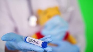 close-up, doctors hand in blue medical glove holds a test tube with inscription coronavirus against background of kids teddy bear in medical mask. Concept of particular danger of coronavirus for