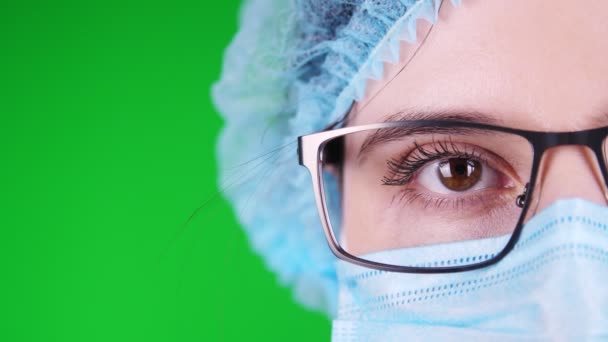 Groene achtergrond. close-up, oog, deel van vrouwelijke arts gezicht in bril, in blauw medisch masker en medische cap. — Stockvideo