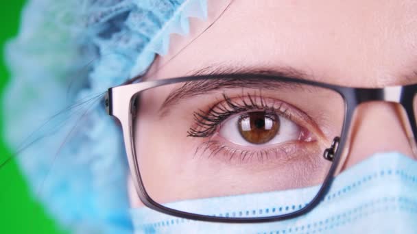 Fundo verde. close-up, olho, parte do rosto médico feminino em óculos, em máscara médica azul e tampa médica . — Vídeo de Stock