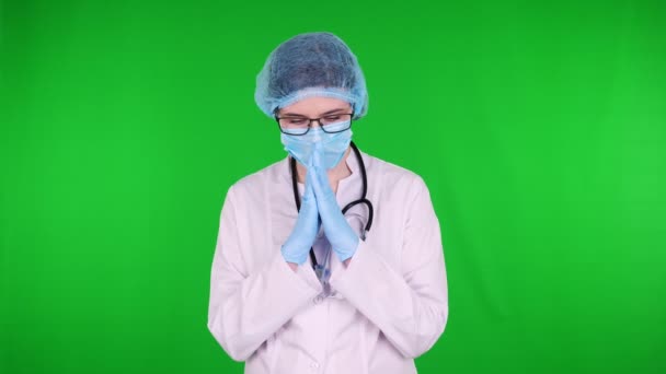 Ritratto di medico in preghiera in occhiali, vestito con uniforme medica bianca, con stetoscopio, con maschera medica blu e cappuccio medico. sfondo verde . — Video Stock