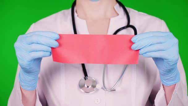 Fondo verde. primer plano, médico vestido con uniforme médico blanco, con un estetoscopio, con las manos en guantes médicos azules está sosteniendo la tarjeta de papel rojo para publicidad, texto o video . — Vídeos de Stock