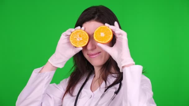 Retrato de una joven doctora, nutricionista vestida con uniforme médico blanco, con estetoscopio, sosteniendo dos mitades de naranja en sus manos, sonriendo. fondo verde . — Vídeo de stock