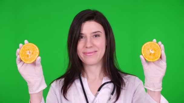 Retrato de una joven doctora, nutricionista vestida con uniforme médico blanco, con estetoscopio, sosteniendo dos mitades de naranja en sus manos, sonriendo. fondo verde . — Vídeo de stock