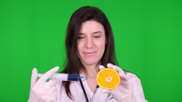 Jeune femme médecin, nutritionniste vêtue d'uniforme médical blanc, avec stéthoscope, injecte avec une seringue un peu de liquide bleu dans la moitié de l'orange. fond vert . — Video