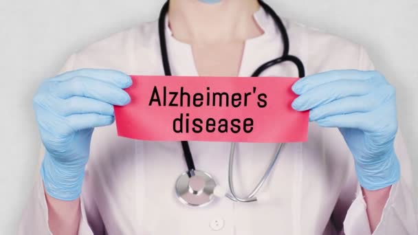Primer plano, las manos en guantes médicos azules sostienen y rasgan la tarjeta de papel rojo con una inscripción enfermedad de Alzheimer. médico vestido con uniforme médico blanco, tiene un estetoscopio . — Vídeos de Stock