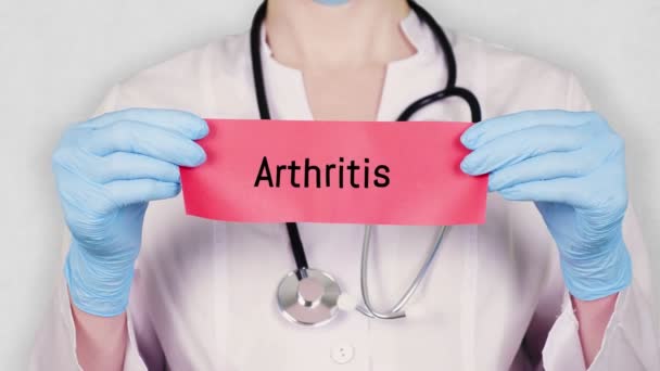 Closeup, hands in blue medical gloves hold and tear red paper card with an inscription arthritis. doctor dressed in white medical uniform, has a stethoscope. — Stock Video