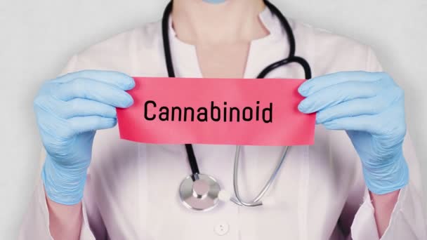 Yakın plan, mavi eldivenli eller kırmızı kağıt kartı Cannabinoid yazısıyla tutuyor ve yırtıyor. Beyaz üniformalı bir doktorun steteskobu var.. — Stok video