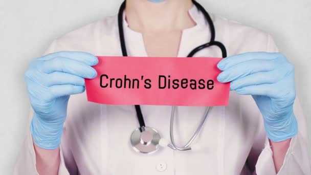 Närbild, händer i blå medicinska handskar hålla och riva rött papper kort med en inskription Crohns sjukdom. läkare klädd i vit medicinsk uniform, har ett stetoskop. — Stockvideo