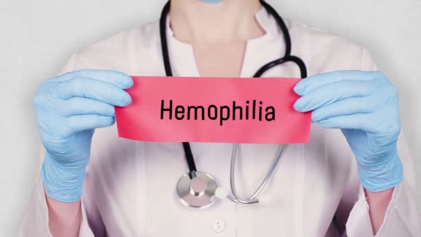 Closeup, hands in blue medical gloves hold and tear red paper card with an inscription Hemophilia. doctor dressed in white medical uniform, has a stethoscope. — Stock Video