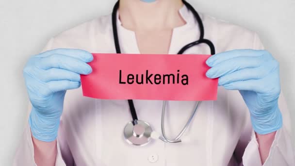 Primer plano, las manos en guantes médicos azules sostienen y rasgan la tarjeta de papel rojo con una inscripción Leucemia. médico vestido con uniforme médico blanco, tiene un estetoscopio . — Vídeo de stock