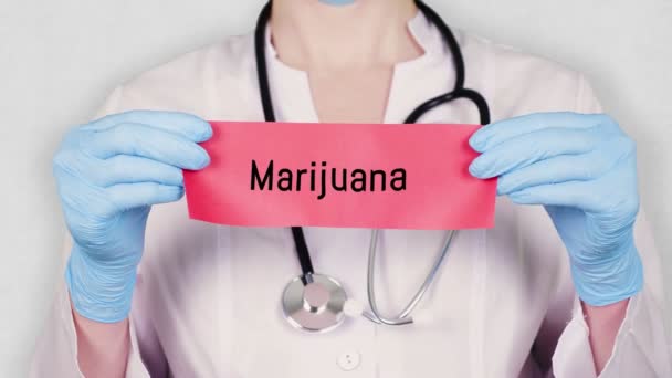 Close-up, handen in blauwe medische handschoenen houden en scheuren rode papieren kaart met een inscriptie marihuana. arts gekleed in wit medisch uniform, heeft een stethoscoop. — Stockvideo