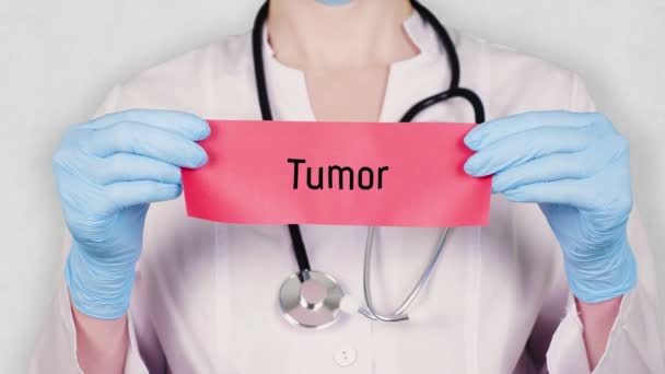 Close-up, mãos em luvas médicas azuis segurar e rasgar cartão de papel vermelho com um tumor inscrição. médico vestido com uniforme médico branco, tem um estetoscópio . — Vídeo de Stock