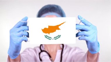 Yakın plan. Gözlüklü doktor, mavi tıbbi şapkalı, eldivenli, Kıbrıs bayrağıyla tıbbi maske takıyor. Doktorlar, koronavirüs sırasında devletin koruması, küresel salgın. Kavram.