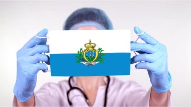 Yakın plan. Gözlüklü doktor, mavi tıbbi şapkalı, eldivenli San Marino bayrağı takan tıbbi maske. Doktorlar, koronavirüs sırasında devletin koruması, küresel salgın. Kavram.