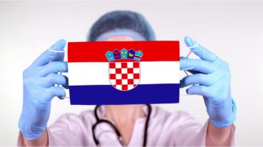 Yakın plan. Gözlüklü doktor, mavi tıbbi başlık, eldivenler Hırvatistan bayrağıyla el maskesi tutuyor. Doktorlar, koronavirüs sırasında devletin koruması, küresel salgın. Kavram.