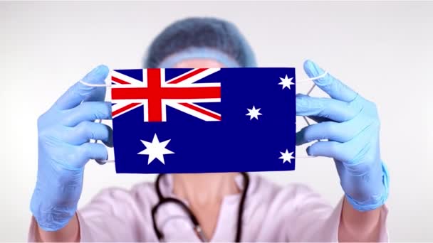 Primo piano. Medico in occhiali, berretto medico blu, guanti tiene in mano maschera medica con bandiera australiana. Cura dei medici, protezione dallo stato durante l'epidemia globale, coronavirus. Concetto . — Video Stock